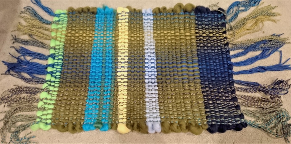 finished weaving sample - a bathmat sized rectangle of chunky cloth. The weft is in stripes of dark navy, olive green, light blue, yellow, turquoise and line green. The selvedges are very bulgy and uneven.