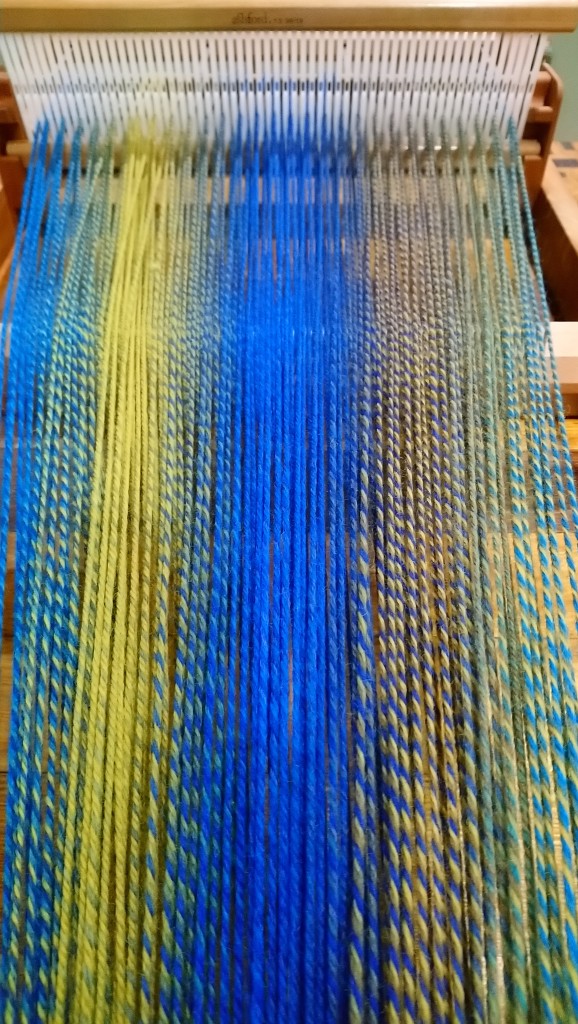 In-progress photo of warping a rigid heddle loom, showing the heddle in the background and the warp spread out toward the camera. The warp yarn is multi-coloured and makes stripes in combinations of blue, green, and yellow.